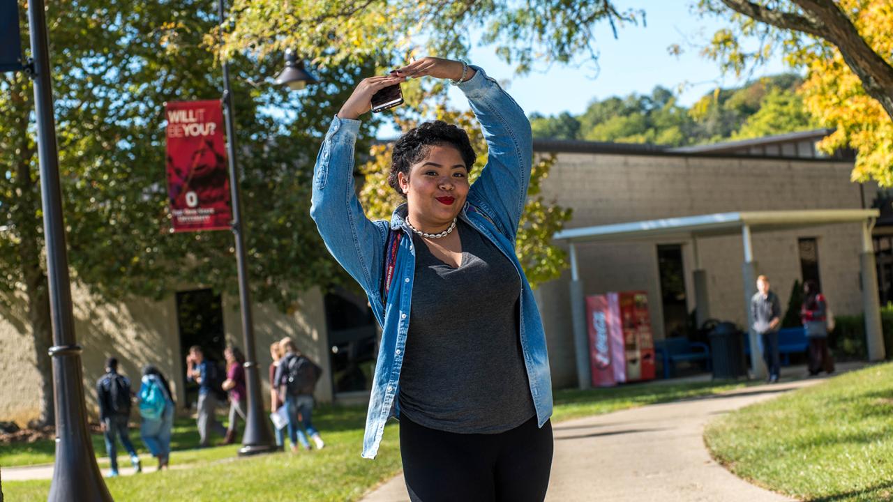 A student stands on campus with her arms formed in the shape of an O above her head.