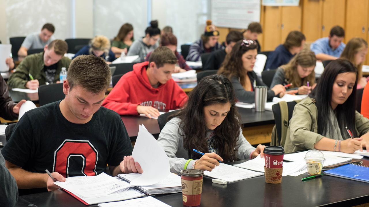 A full class of students sits at desks with their heads downward looking at a paper test. 