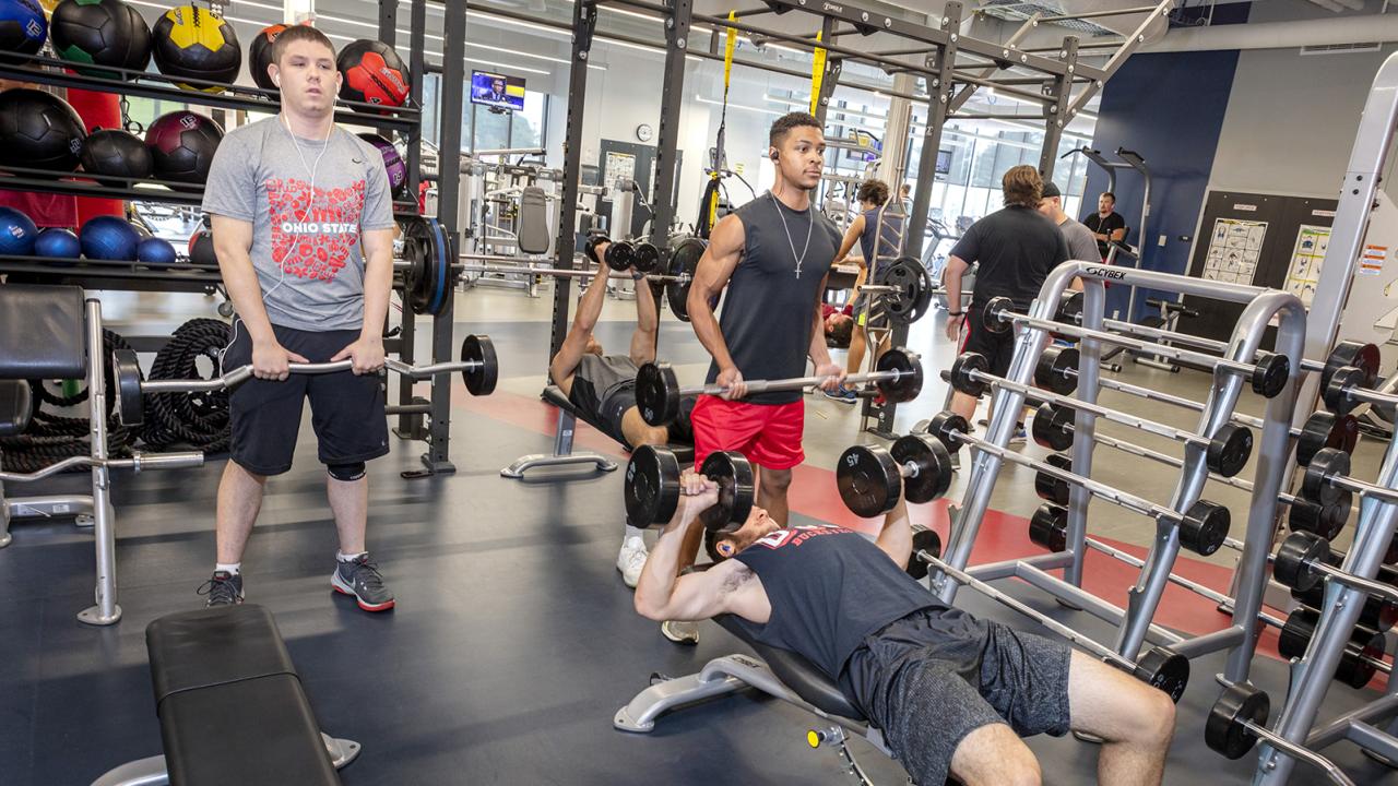 Two students hold use a barbell while a third student lies on a bench pressing dumbells.
