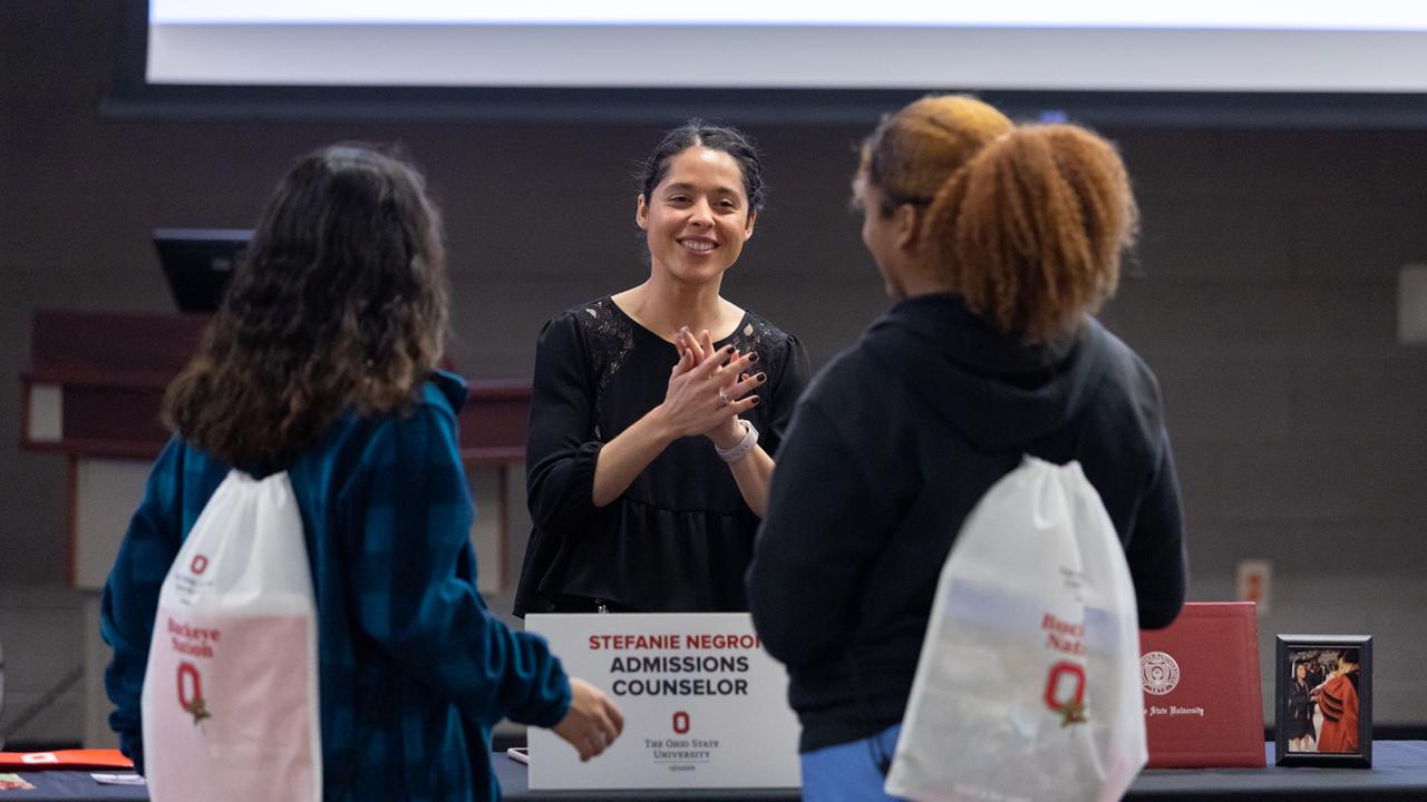 Ad admissions representative converses with her hands in motion with two students who have their backs to the camera. 