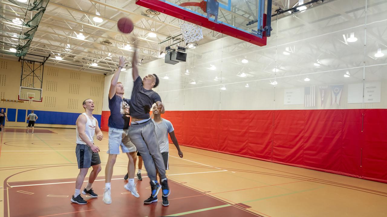 A student blocks the shot of another student playing basketball while teams stand by. 