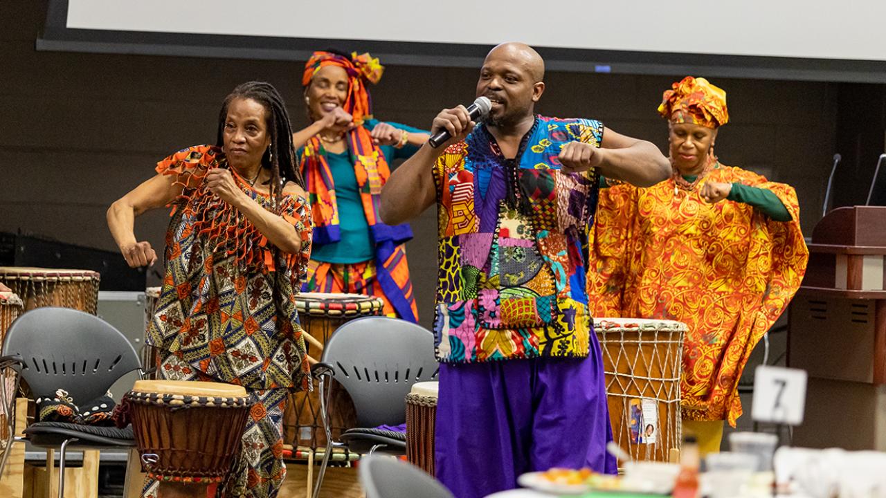 A group of musicians in brightly colored African apparel dance while singing and drumming.