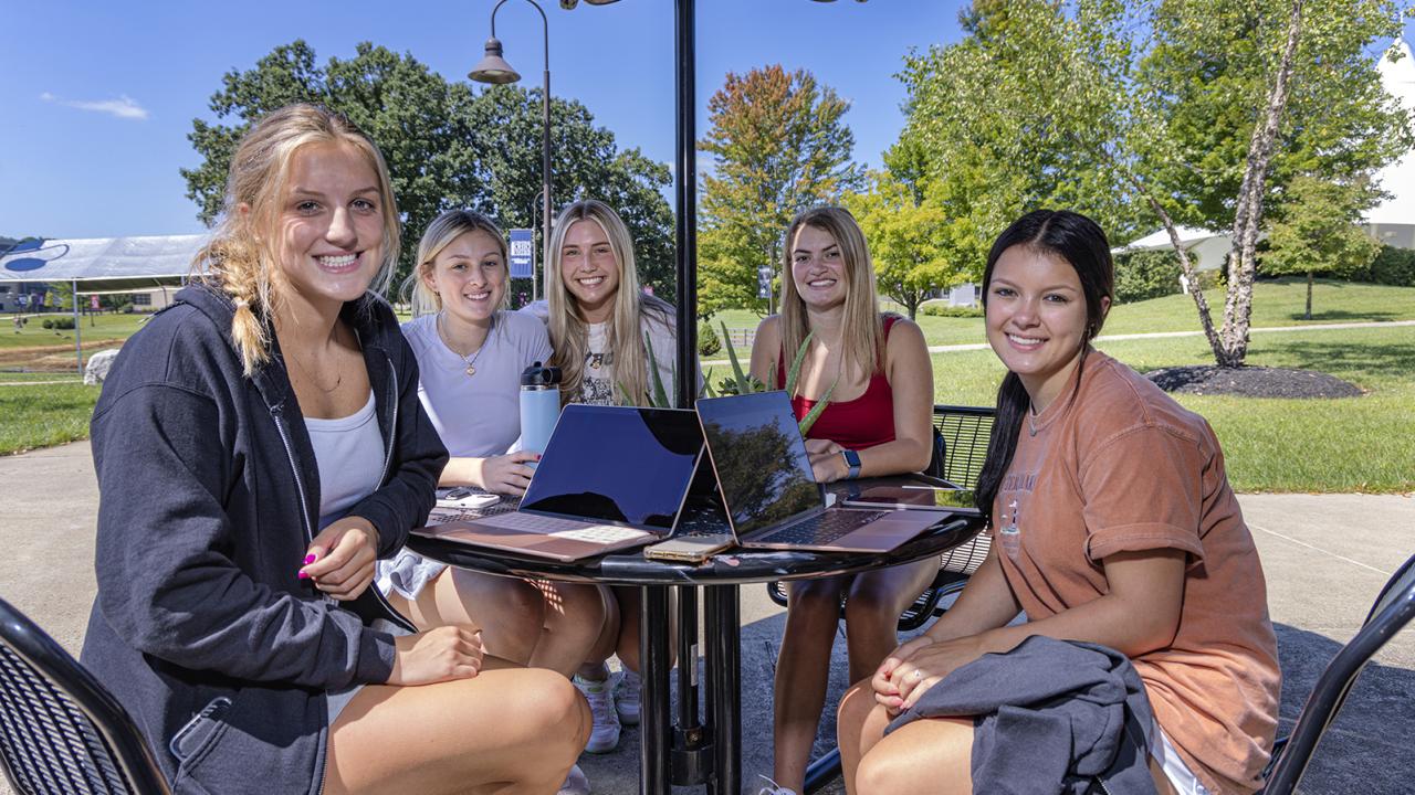 Five female students sit at a table outside on campus.