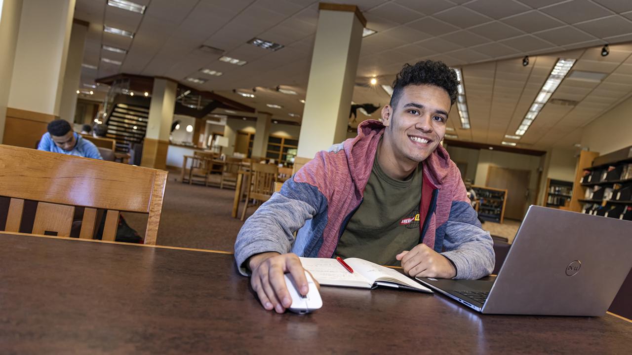 A male student in the library looks up from his laptop smiling while his hands rests on a mouse.