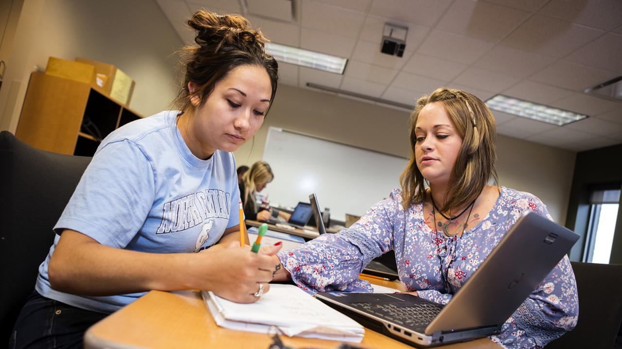 A student leans over a desk to write in a notebook of another student seated next to her.