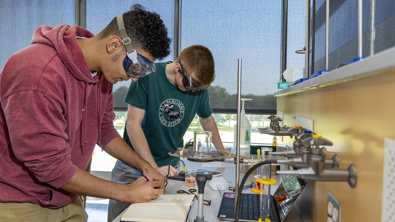 Two male students wearing goggles lean over a table to write in a notebook on a table with beakers and lab equipment.