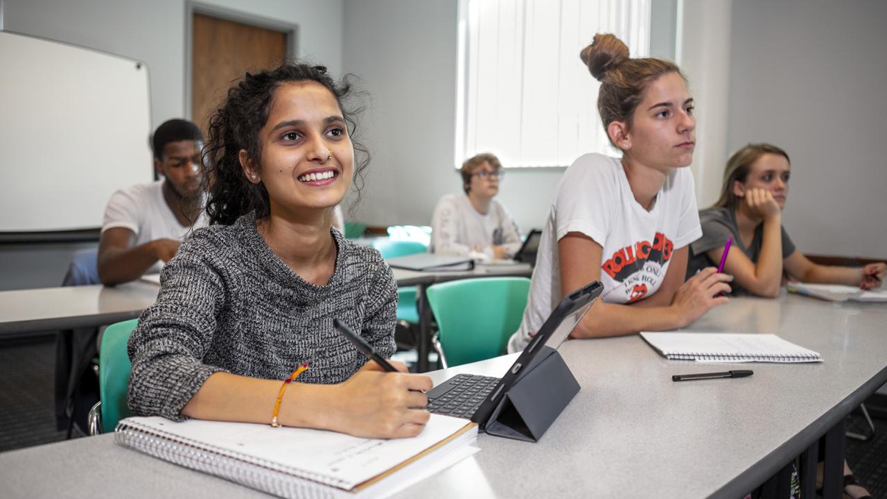 In a classroom of students, a smiling female student looks up to the front of the room writing in a notebook with an iPad open beside her.