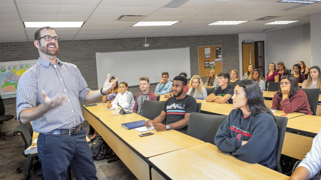A professor stands with his hands open in conversation at the front of a classroom.
