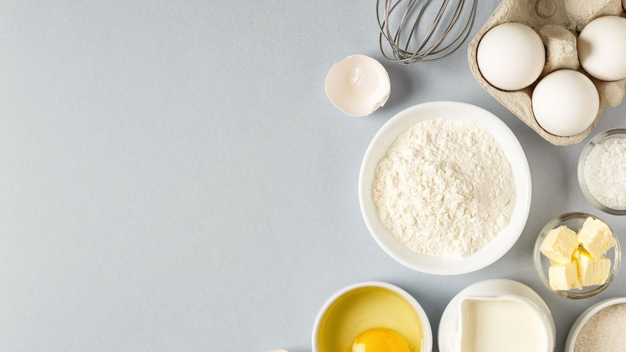 A whisk, carton of eggs, flower, butter, and other food ingredients.