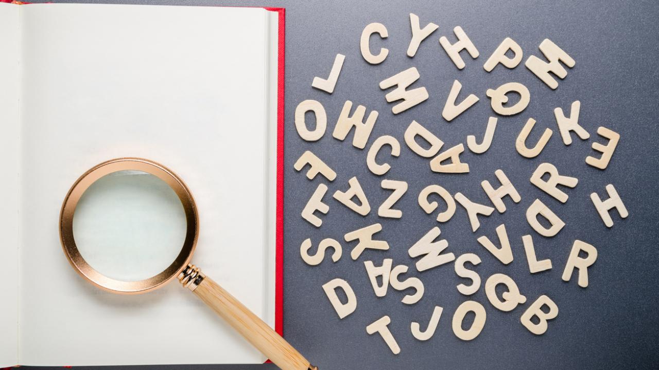 A magnifying glass sits on a blank page of an open book next to scattered alphabet letters. 