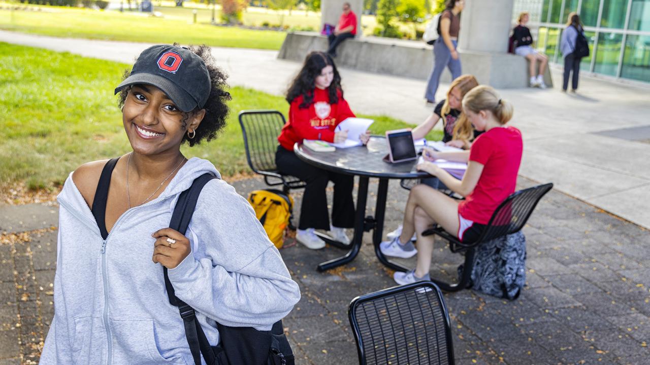 A female student stands smiling while a group of students is seated at an outdoor table behind her.