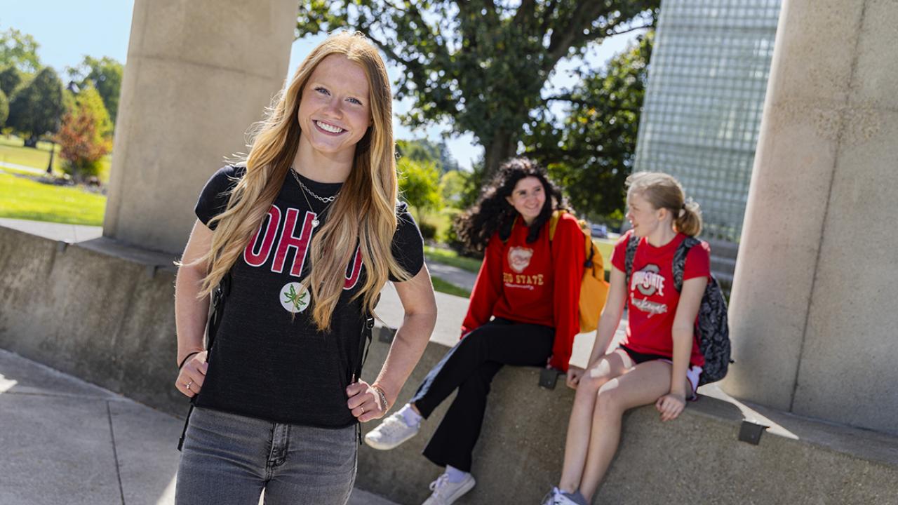 A female student stands smiling while a group of students is seated at an outdoor table behind her.