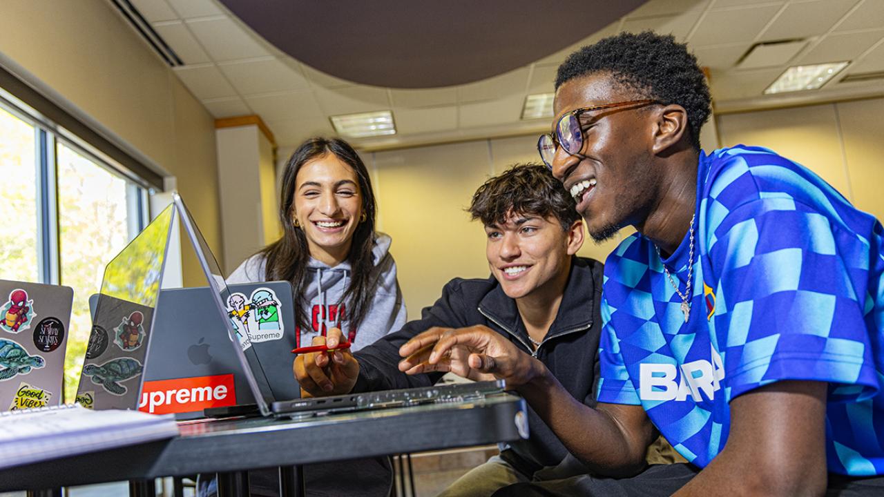 Three students sit around a table looking at a laptop and smiling.