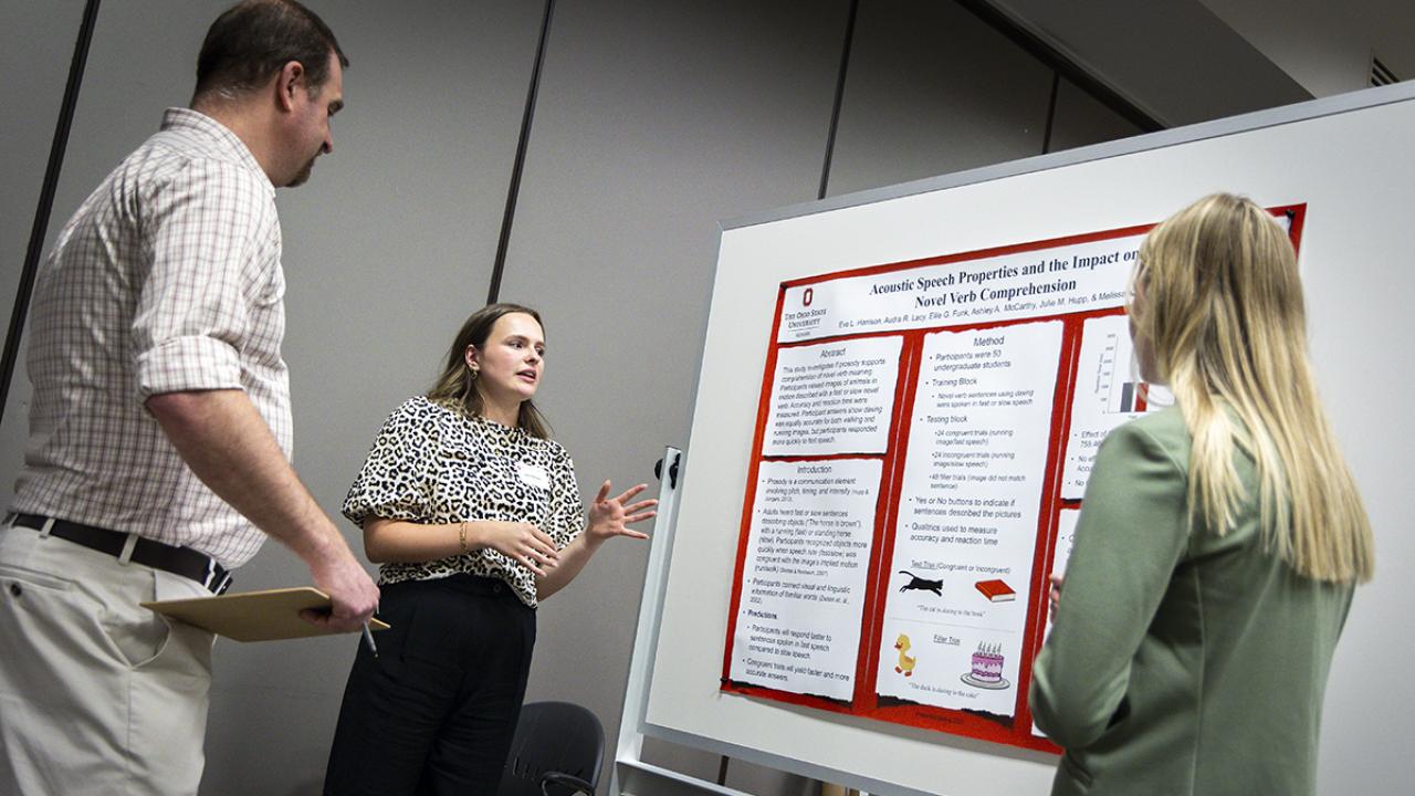 A student stands next to her poster presentation talking to a spectator. 