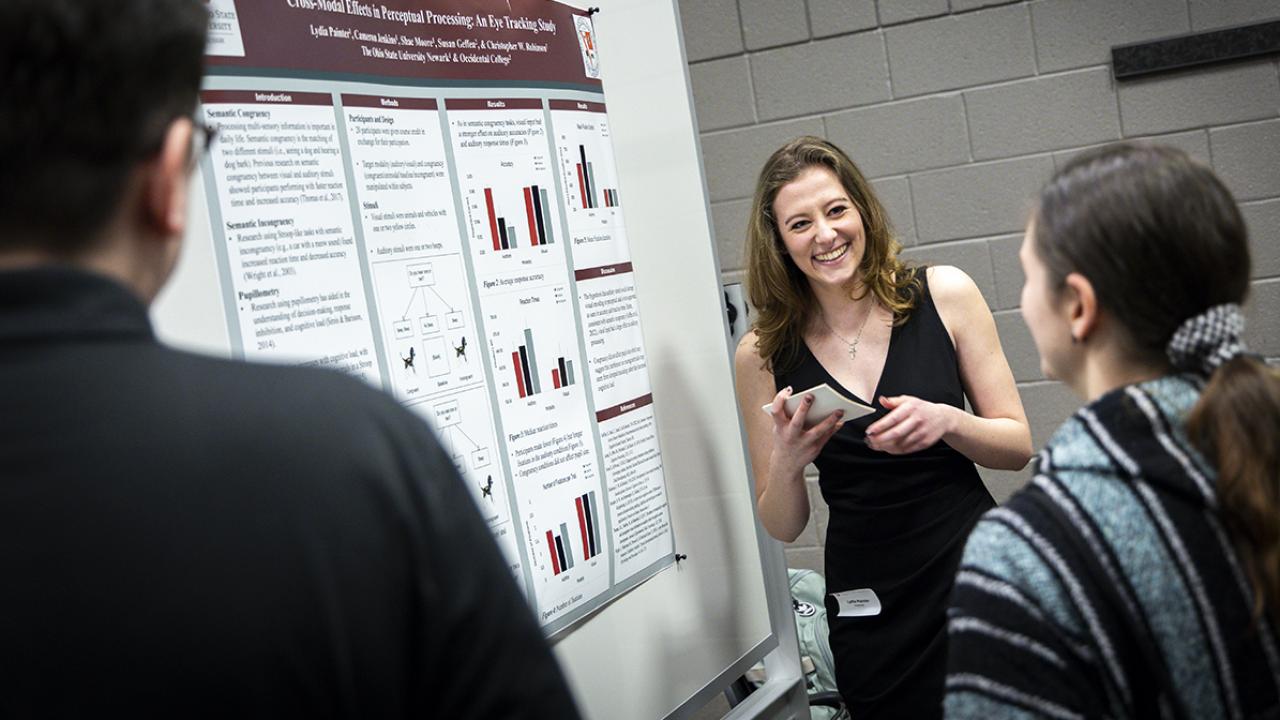 A student smiles at an onlooker while sharing her research poster. 
