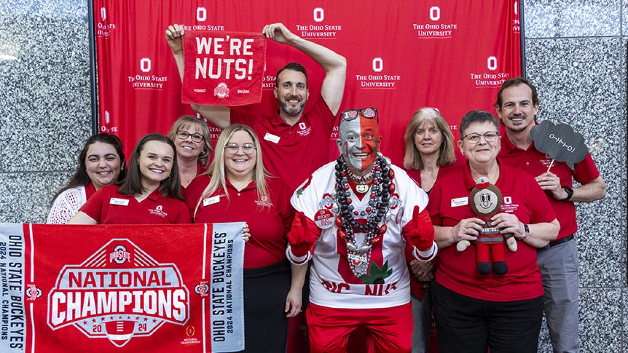 Academic advising staff members hold Buckeye-themed signs and objects as they pose with Ohio State superfan Big Nut.