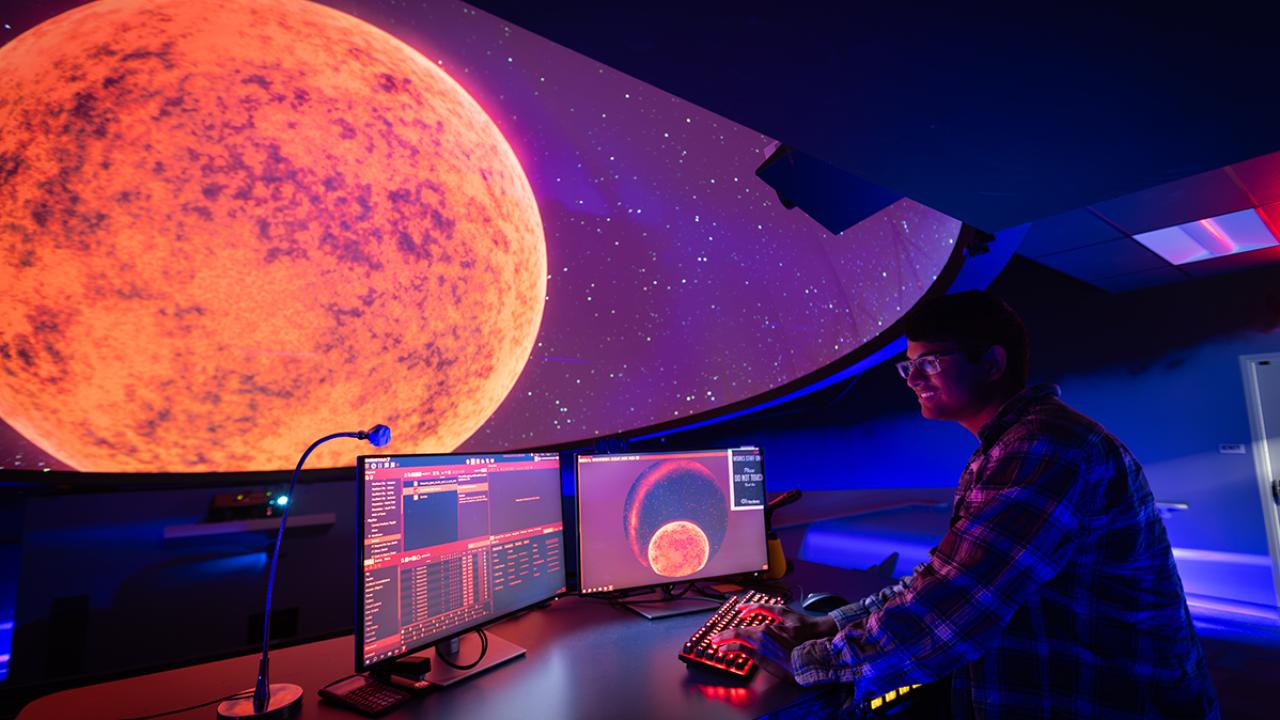 A person inside a dark planetarium stands at a computer selecting the display on the planetarium's theater screen.