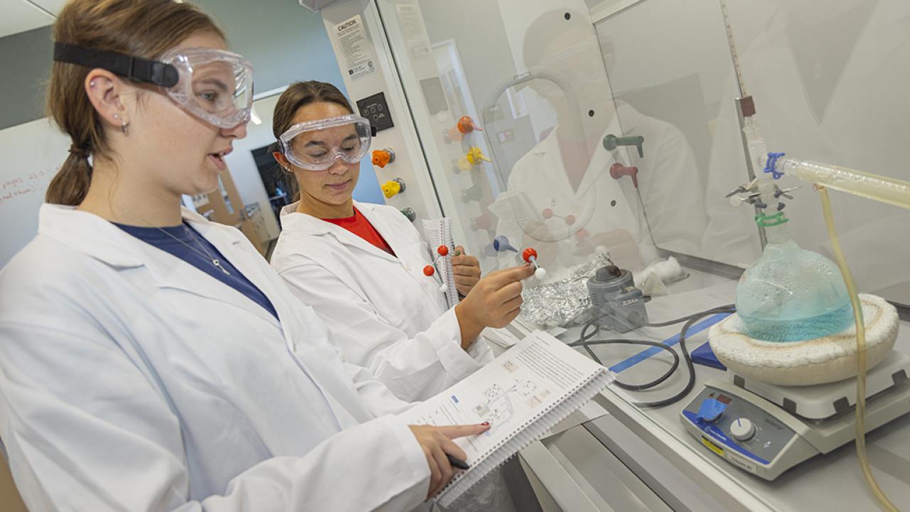 One student wearing a white lab coats and safety goggles points to an image in a book while a second student in a lab coat and safety goggles holding molecular figures looks at the book.