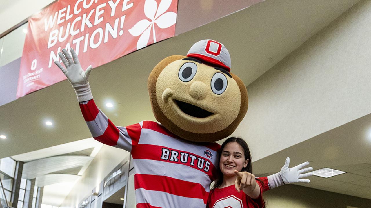 In front of a sign that says "Welcome to Buckeye Nation," Brutus the Buckeye mascot stands with his arms outstretched next to a student who is pointing toward the camera. 