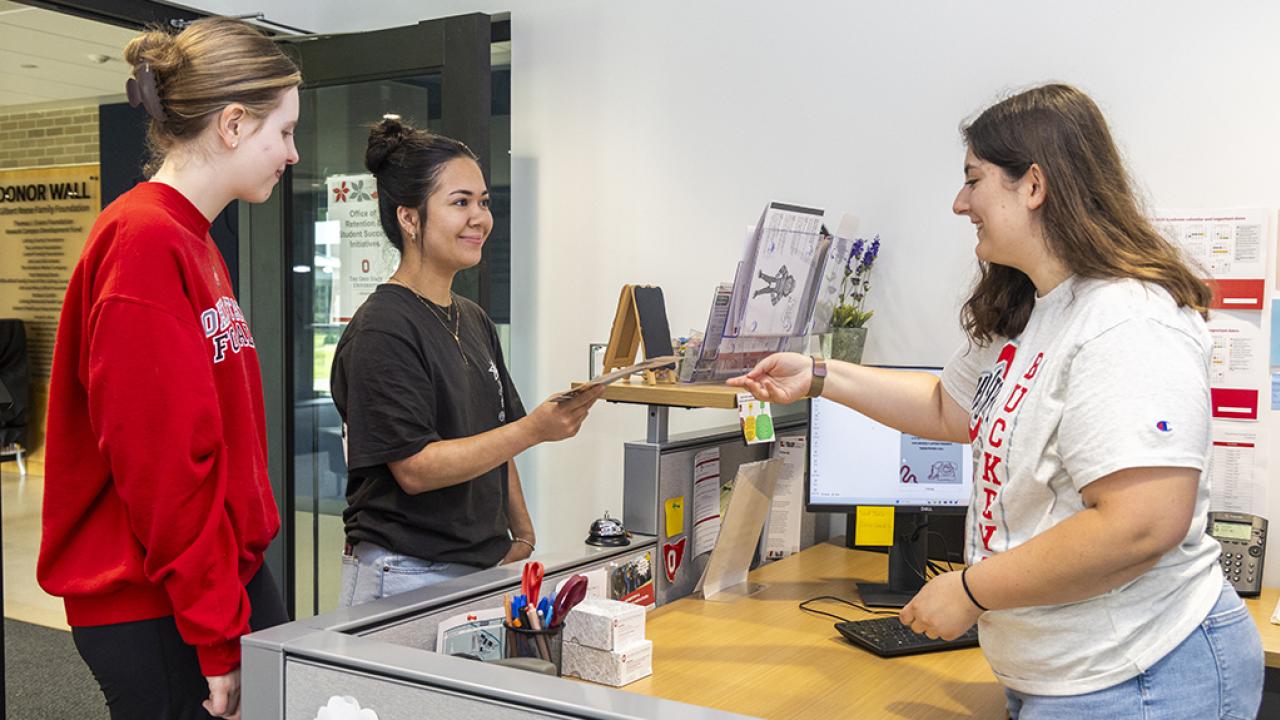 A student employee working the front desk stands up behind her desk to give a brochure to a student visitor while another student stands waiting.