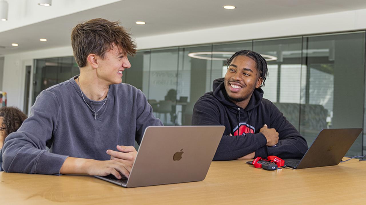 Two students seated at a table with open laptops look toward each other with smiles on their faces.